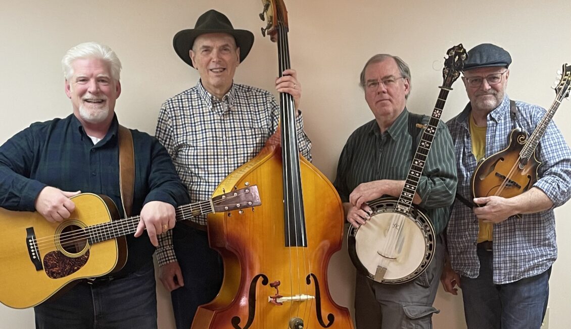 A posed photograph of the four members of the High Peaks Bluegrass band standing infront of an ivory colored wall