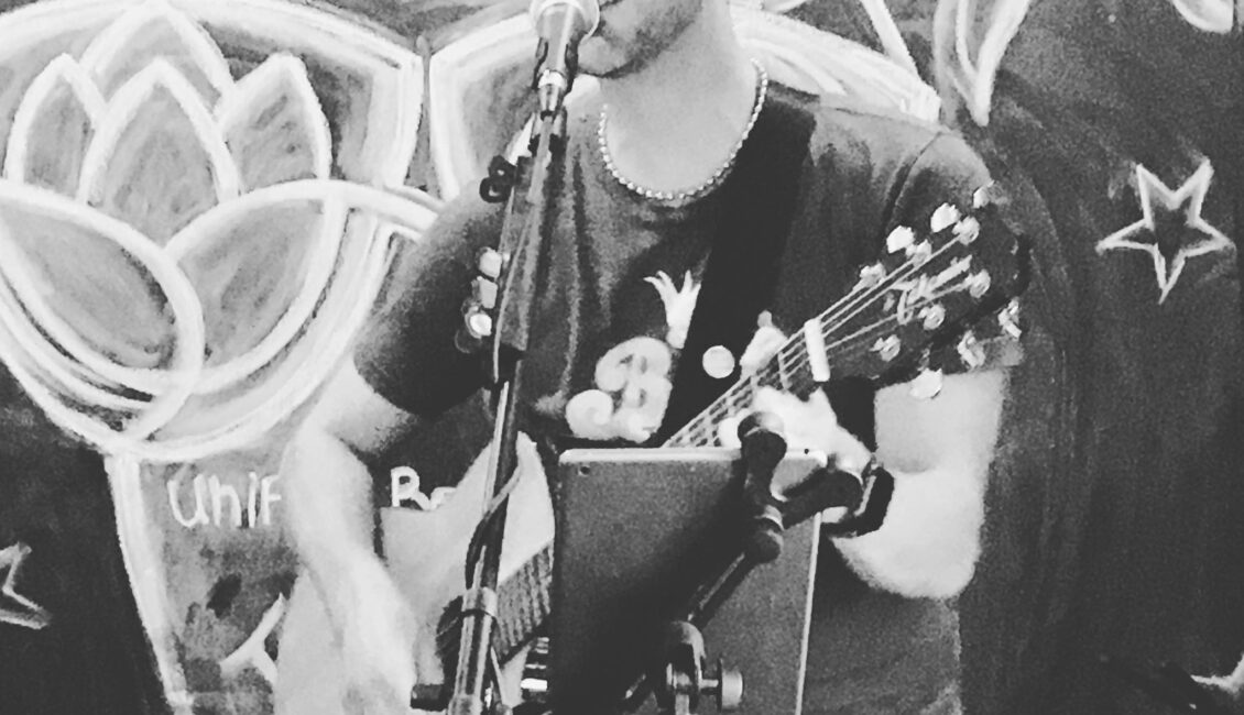 Black and white photograph of acoustic musician Mike Grosshandler playing his guitar while singing passtionately outdoors in front of a mural.
